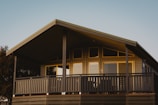 A modern, single-story house with a peaked roof and several windows. The front of the house features a spacious porch with wooden railing painted in dark hues. The exterior walls are a light shade, contrasting with the dark roof and railings. Tall trees can be seen in the background under a clear blue sky.