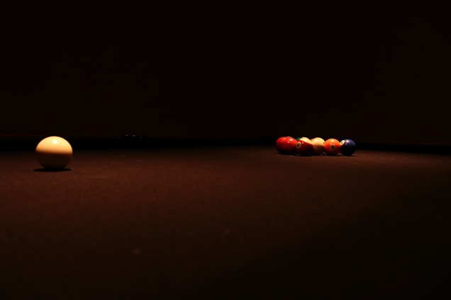 A dimly lit billiard table features a single white cue ball positioned on the left side with a group of colored billiard balls lined up neatly on the right. The focus is on the contrast between the solitary cue ball and the clustered colored balls, with shadows creating an ambiance of anticipation and calm.