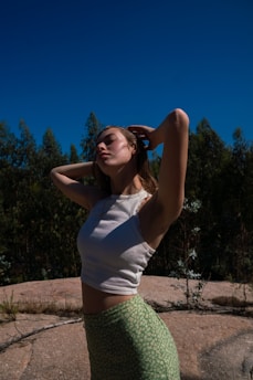 A person stands outdoors with eyes closed and arms raised, surrounded by lush greenery under a clear blue sky. The lighting creates soft shadows and highlights.