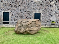 A large, sculpted stone sits on a grassy area in front of a building with a detailed stone facade. The wall features a pattern of stones, creating a textured appearance. Two dark wooden doors are visible on the building, framed by rectangular stone blocks. A small potted plant is placed near one of the doors.