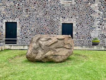 A large, sculpted stone sits on a grassy area in front of a building with a detailed stone facade. The wall features a pattern of stones, creating a textured appearance. Two dark wooden doors are visible on the building, framed by rectangular stone blocks. A small potted plant is placed near one of the doors.