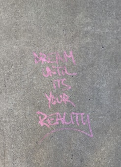 Pink chalk writing on a concrete pavement reads 'Dream until it's your reality.' The text appears artistic and slightly uneven against the rough texture of the sidewalk.