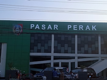 Facade of a building with the name 'Pasar Perak' displayed on a green signage. Below the sign, there are multiple rows of black-tinted windows. Several vehicles are parked in front, with a few individuals standing nearby. Electric cables run across the image, partially obscuring the view.