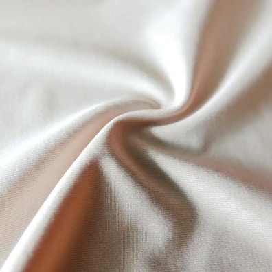 A close-up of a piece of fabric with soft folds and a smooth texture. The material appears to be light-colored, possibly beige or cream, catching the light at various angles and creating a subtle play of light and shadow.