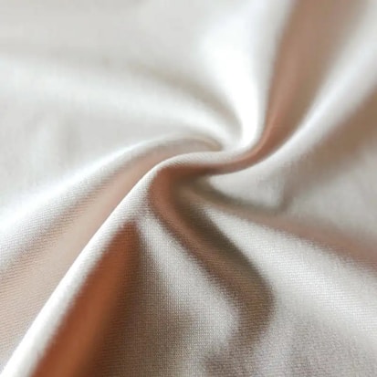 A close-up of a piece of fabric with soft folds and a smooth texture. The material appears to be light-colored, possibly beige or cream, catching the light at various angles and creating a subtle play of light and shadow.