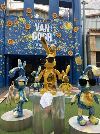 A group of colorful rabbit sculptures, each adorned with various patterns reminiscent of Van Gogh's art, stand on reflective pedestals. The large backdrop features the text 'Van Gogh: The Immersive Experience' with a starry night and sunflower motif. Some of the rabbit sculptures are wearing sunglasses, adding a playful element to the overall artistic installation.