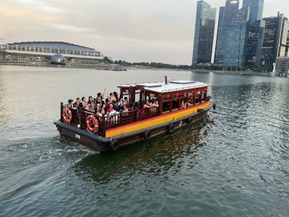 A small wooden tour boat, filled with people, is navigating through a body of water. Modern skyscrapers rise in the background, and a large, illuminated stadium structure is visible to the left on the shoreline.
