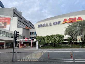 A large shopping mall with a sign that reads 'MALL OF ASIA' in prominent letters. The building is modern with a gray facade, and there are palm trees and plants in front. A festive archway with ‘HAPPY HOLIDAYS 2022’ decorates the entrance, with candy cane stripes. There's a traffic light and a few barriers marking the pedestrian area.