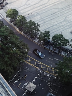 Aerial view of a street with a black car driving along a curved road bordered by trees and a large tiled plaza. Several people are walking on the pavement and crossing areas are marked with yellow lines. The scene is urban with additional structures such as an overhang and various parked motorbikes.