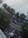 Aerial view of a street with a black car driving along a curved road bordered by trees and a large tiled plaza. Several people are walking on the pavement and crossing areas are marked with yellow lines. The scene is urban with additional structures such as an overhang and various parked motorbikes.