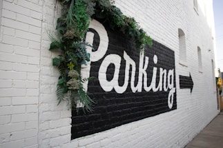 A brick wall painted white features a large black rectangle with the word 'Parking' in bold white letters. There is a directional arrow pointing right. Green foliage decorates the top left corner of the signage.