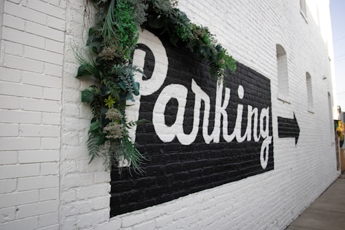 A brick wall painted white features a large black rectangle with the word 'Parking' in bold white letters. There is a directional arrow pointing right. Green foliage decorates the top left corner of the signage.