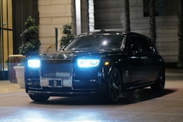 A luxury black car with a prominent grille and bright headlights parked in an upscale urban setting. The surrounding environment features potted plants and elegant architectural details.