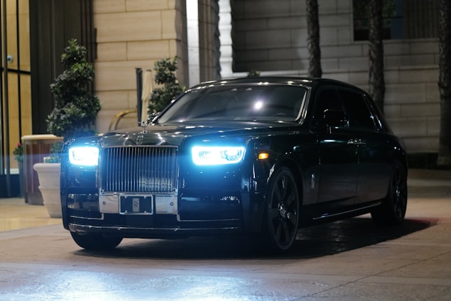 A luxury black car with a prominent grille and bright headlights parked in an upscale urban setting. The surrounding environment features potted plants and elegant architectural details.