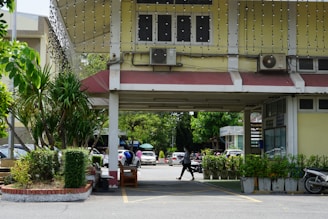 A building entrance with a covered walkway, featuring potted plants and decorative string lights overhead. Air conditioning units are mounted above the entrance. A person is walking through the area, and several vehicles are parked nearby, including cars and motorcycles. The surrounding area is lush with greenery.