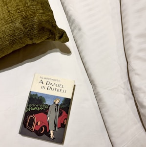 A book titled 'A Damsel in Distress' by P.G. Wodehouse lies on a white bed sheet. The cover illustration features a woman in a long coat standing beside a red vintage car, set against a backdrop of greenery. A textured green pillow is positioned to the left.
