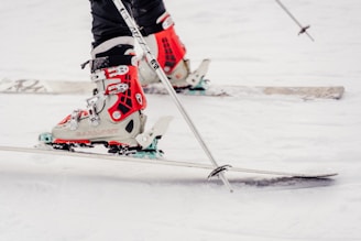 A close-up of a person wearing ski boots attached to skis on a snowy ground. The boots are primarily red with white and black accents, and the skis are lined with snow. The skier is holding ski poles.