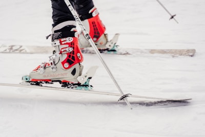 A close-up of a person wearing ski boots attached to skis on a snowy ground. The boots are primarily red with white and black accents, and the skis are lined with snow. The skier is holding ski poles.