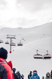 Skiers gather near a ski lift on a snowy mountain slope. The sky is overcast, and there are several chairlifts visible, transporting people across the scenic, snow-covered landscape. People are dressed in winter gear, preparing for skiing.