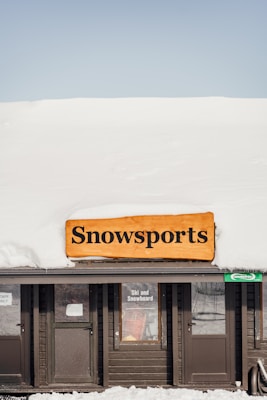 A wooden building with a thick layer of snow on the roof displays a sign reading 'Snowsports' in bold black letters. Below the sign, in the window, another sign advertises 'Ski and Snowboard'. The scene conveys a winter setting and suggests a facility for winter sports activities.