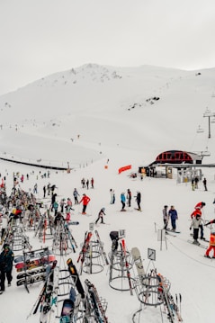 A snowy ski resort with numerous people engaging in skiing activities. Various colorful skis and snowboards are organized on racks in the foreground. Skiers, dressed in winter gear, are scattered across the snow-covered landscape. A mountain slope looms in the background, with a ski lift visible on the right and a small building nearby.