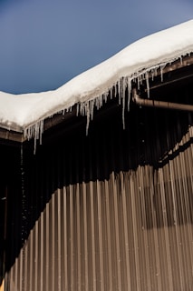 A sloped roof covered with a thick layer of snow, with icicles hanging from the edge. The building has a corrugated metal wall creating a distinct shadow pattern.