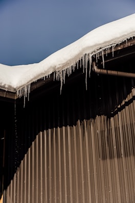 A sloped roof covered with a thick layer of snow, with icicles hanging from the edge. The building has a corrugated metal wall creating a distinct shadow pattern.