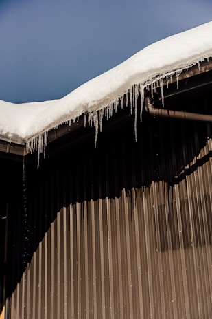 A sloped roof covered with a thick layer of snow, with icicles hanging from the edge. The building has a corrugated metal wall creating a distinct shadow pattern.