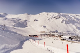 A snowy mountain landscape with a ski resort at the base. Numerous skiers and snowboarders are visible on the slopes and near the resort buildings. The scene includes several buildings with snow-covered roofs, a ski lift, and red poles marking a path on the snow.