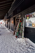 A group of skis and ski poles are propped up against the exterior of a building on a snow-covered path. The building has dark walls and several posters and signs on them. Snow is piled up against the side of the building.