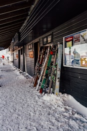 A group of skis and ski poles are propped up against the exterior of a building on a snow-covered path. The building has dark walls and several posters and signs on them. Snow is piled up against the side of the building.