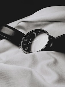 A classic analog wristwatch with a dark leather strap lies on a textured white fabric. The watch face is black with minimalist numbering, reflecting a simple yet elegant design.