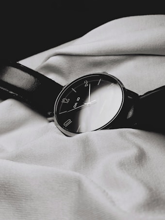 A classic analog wristwatch with a dark leather strap lies on a textured white fabric. The watch face is black with minimalist numbering, reflecting a simple yet elegant design.