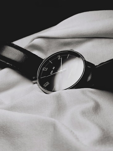 A classic analog wristwatch with a dark leather strap lies on a textured white fabric. The watch face is black with minimalist numbering, reflecting a simple yet elegant design.
