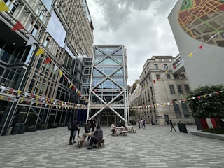 An outdoor urban courtyard surrounded by modern and traditional architecture. The space features glass-fronted buildings with crisscrossing metal beams and colorful triangular bunting strung across the courtyard. People are gathered around wooden picnic tables, and a large mural is visible on one building wall.