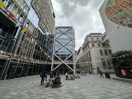 An outdoor urban courtyard surrounded by modern and traditional architecture. The space features glass-fronted buildings with crisscrossing metal beams and colorful triangular bunting strung across the courtyard. People are gathered around wooden picnic tables, and a large mural is visible on one building wall.