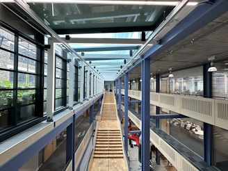 A modern, spacious interior with a high ceiling, featuring a long wooden staircase leading down the center. Glass walls line the corridor on the left side, providing a view of green plants outside. The right side has open office spaces with people working at desks. Overhead, large steel beams and hanging industrial-style lights add to the contemporary design.