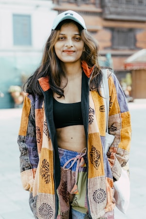 A young woman with long hair wears a colorful patchwork jacket, a black crop top, and patterned shorts. She has a light blue cap and carries a white bag on her shoulder. The background features a blurred urban setting with a mix of traditional and modern architectural elements.