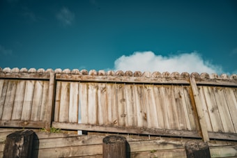 A rustic wooden fence with vertical planks and a slightly weathered texture, set against a bright blue sky with scattered white clouds.
