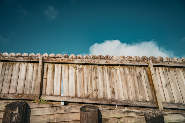 A rustic wooden fence with vertical planks and a slightly weathered texture, set against a bright blue sky with scattered white clouds.