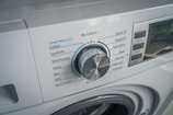 A close-up view of a washing machine control panel with various washing options such as Speed Wash, Cotton, Synthetic, and others. The dial knob is prominently positioned, slightly tilted to the left. Adjacent buttons display functions like Delay and Temperature.