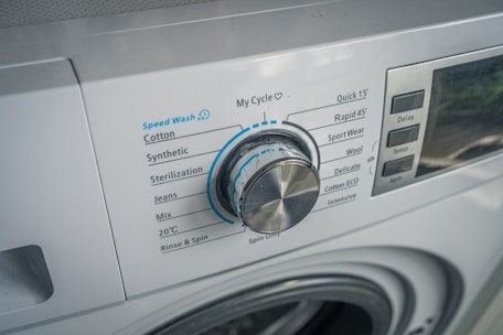 A close-up view of a washing machine control panel with various washing options such as Speed Wash, Cotton, Synthetic, and others. The dial knob is prominently positioned, slightly tilted to the left. Adjacent buttons display functions like Delay and Temperature.