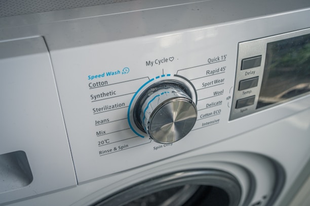 A close-up view of a washing machine control panel with various washing options such as Speed Wash, Cotton, Synthetic, and others. The dial knob is prominently positioned, slightly tilted to the left. Adjacent buttons display functions like Delay and Temperature.