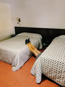 A room with two single beds covered with patterned bedspreads. There is a person lying horizontally across the beds, with only their legs visible wearing yellow stockings and black shoes. The room has a white wall, a wall-mounted lamp, and tiled floor.