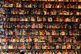 A large collection of various bottles, many containing different types of alcoholic beverages, is displayed on shelves. The bottles are arranged in rows, filling the entire frame with a myriad of colors and labels. The shelves are well-lit, highlighting the colorful and diverse assortment of bottles.