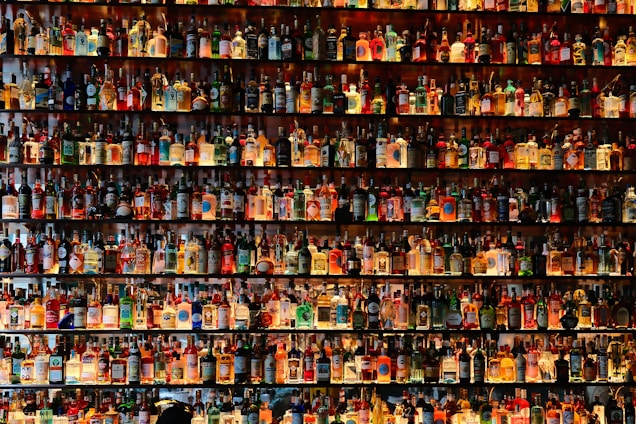 A large collection of various bottles, many containing different types of alcoholic beverages, is displayed on shelves. The bottles are arranged in rows, filling the entire frame with a myriad of colors and labels. The shelves are well-lit, highlighting the colorful and diverse assortment of bottles.