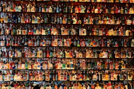 A large collection of various bottles, many containing different types of alcoholic beverages, is displayed on shelves. The bottles are arranged in rows, filling the entire frame with a myriad of colors and labels. The shelves are well-lit, highlighting the colorful and diverse assortment of bottles.