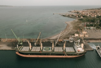 Aerial view of a large cargo ship docked at an industrial port. Multiple cranes along the dock are positioned beside the ship, indicating loading or unloading activities. The calm sea stretches into the distance with a small sailboat visible. In the background, a coastal town with clustered buildings and a narrow strip of sandy shoreline can be seen.