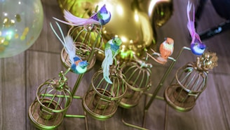 Several colorful artificial birds are perched on ornate, small golden cages on a metal stand. A translucent balloon with gold confetti can be seen in the background, along with a reflective surface casting a glowing light.