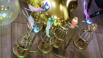 Several colorful artificial birds are perched on ornate, small golden cages on a metal stand. A translucent balloon with gold confetti can be seen in the background, along with a reflective surface casting a glowing light.